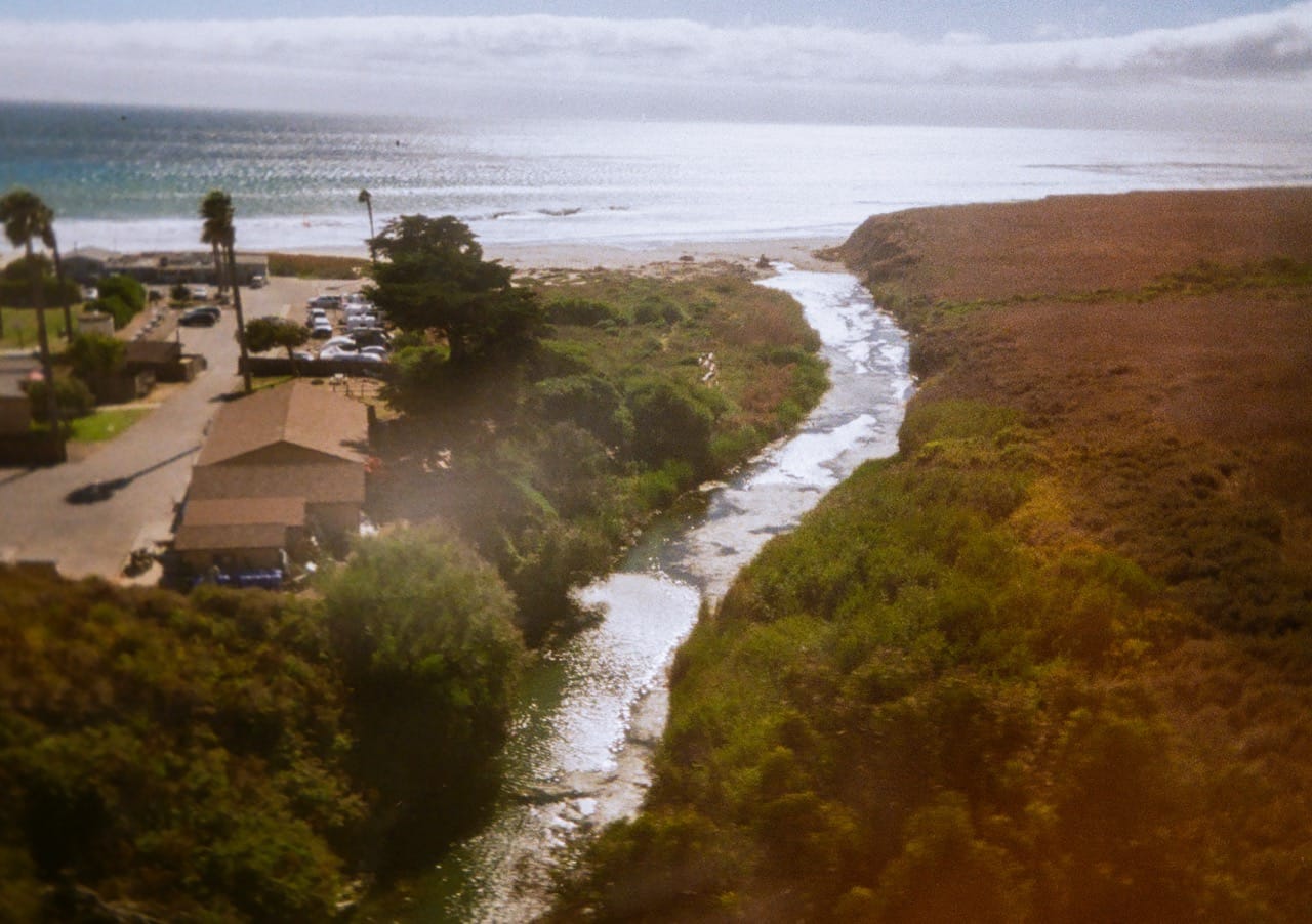 a color photograph of a river leading to the Pacific Ocean with the sun reflecting off both. An open field is on the right side of the river, a parking lot and building on the other