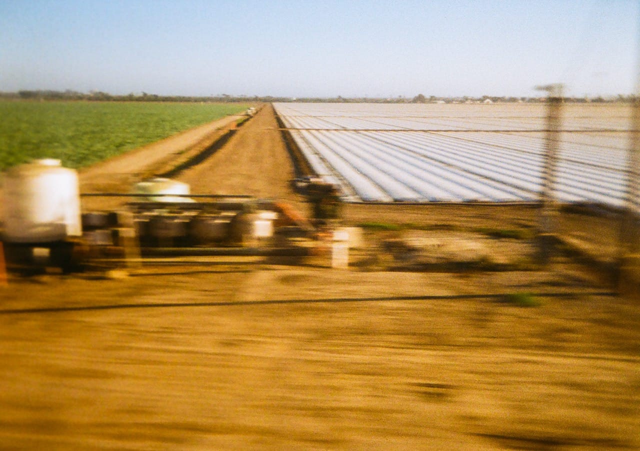 a color photo of agricultural fields stretching into the distance, with a blurry set of industrial tanks in the foreground