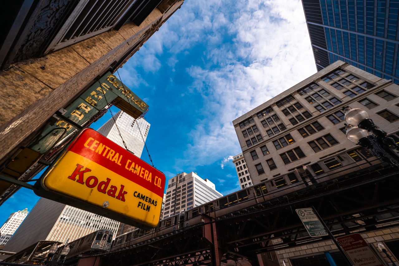 a low-angle color photograph taken in downtown Chicago. An old yellow and red sign in the left foreground reads 'Central Camera Co. Kodak Cameras Film' The El runs behind it and various buildings rise around a cloudy blue sky in the background
