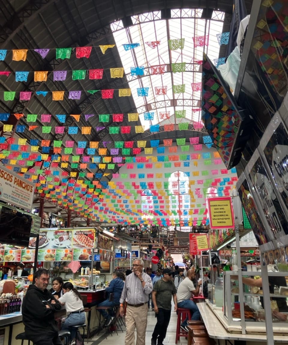 a color photo of the interior of Mercado Hidalgo. Colorful paper flags are strung across the aisle between food vendors. The curved steel roof and skylights in the background