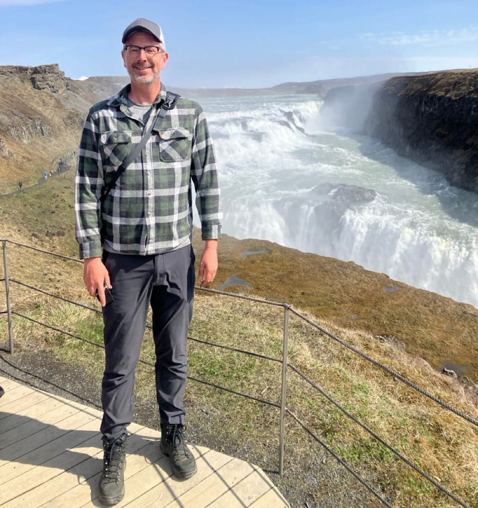 A color photo of J. Mark standing on an observation platform above Gullfoss, Iceland. It's a sunny day and the waterfall rages behind him.