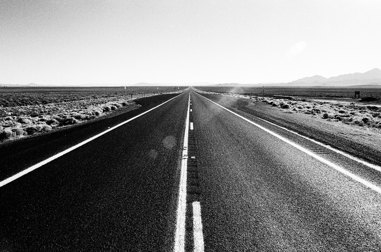 a black and white image of 'The Loneliest Road in America' showing a two-lane highway stretching off through the desert of Nevada to the horizon