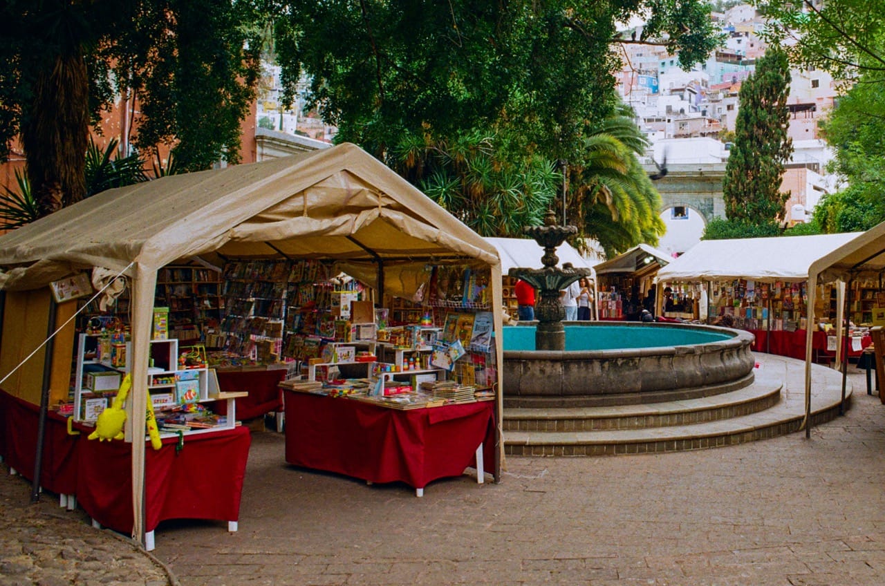a color photo of several tents selling books on a paved plaza. a fountain sits in the center, the city of Guanajuato rises through the trees in the background