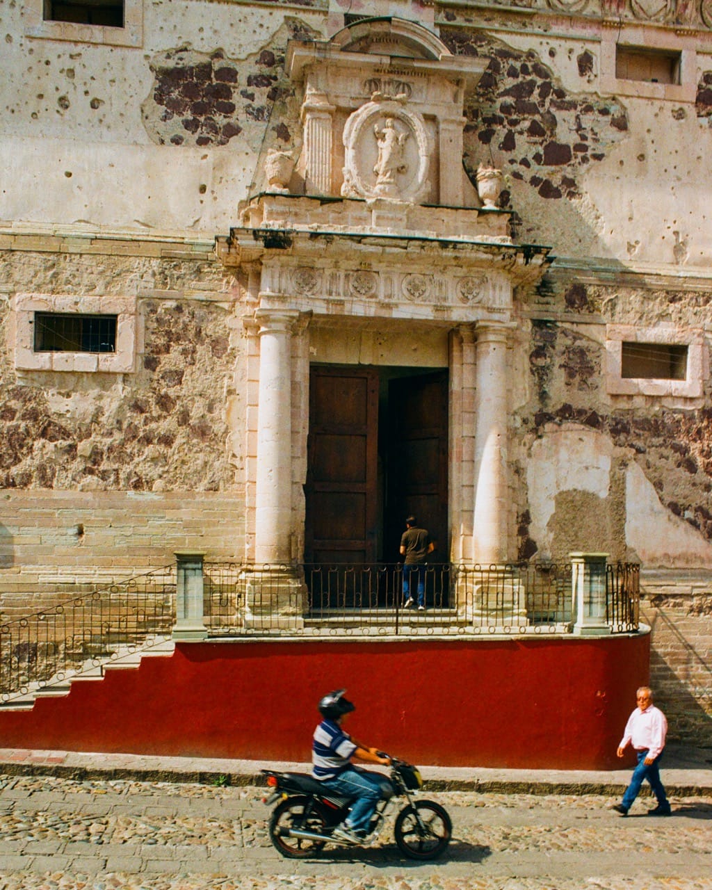 A color photograph of the entry to the Alhóndiga. A man walks through the open wooden door, a second man walks past on the street, a third man crosses on a motorcycle.