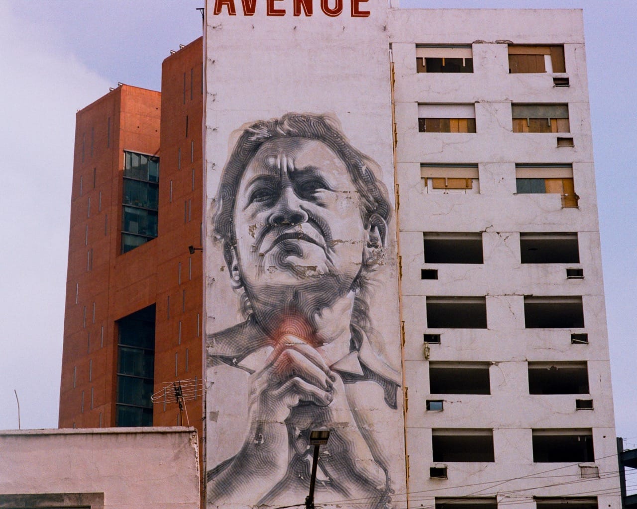 a color photograph of tall buildings in Mexico City. The building in the center has a large mural of a woman clasping her hands together.