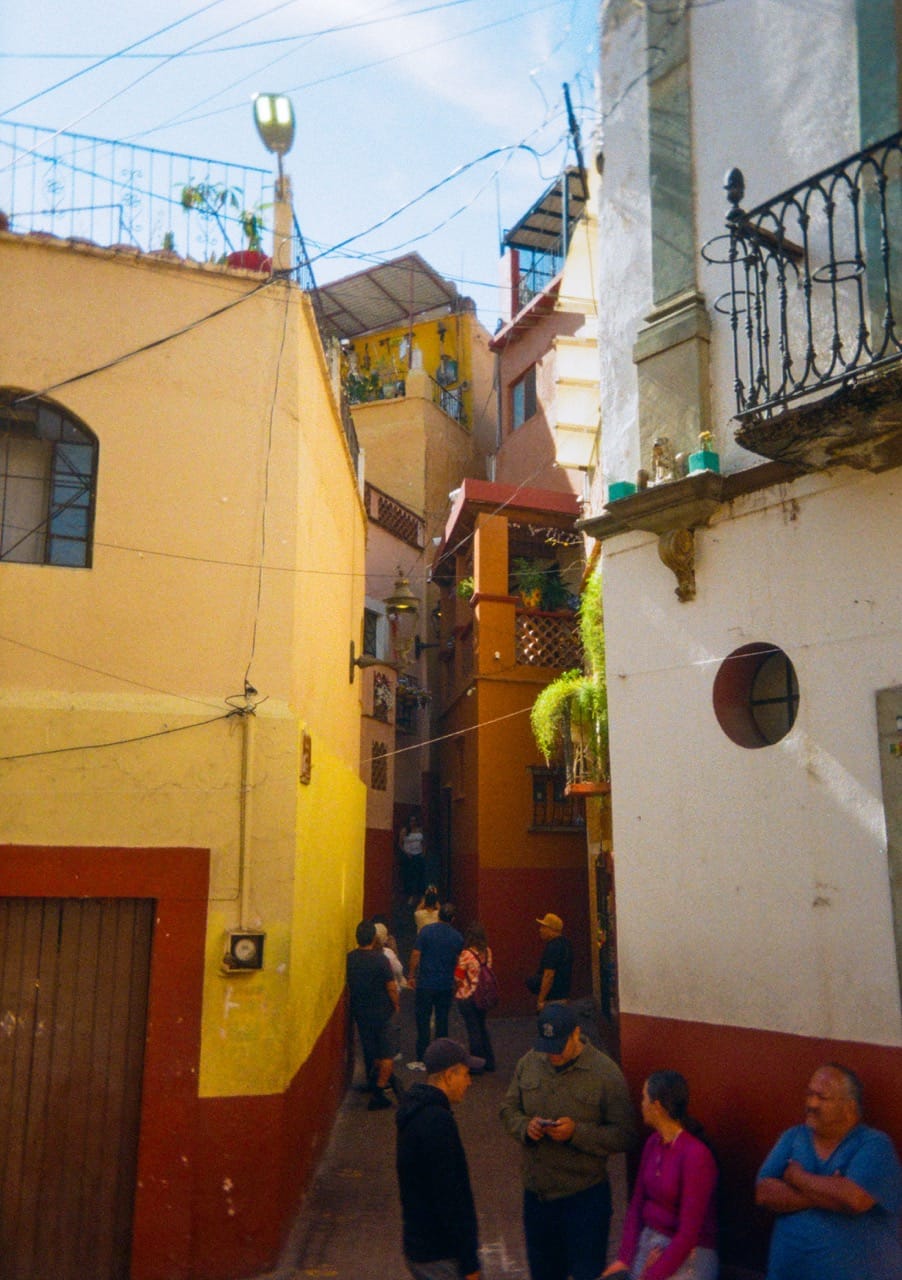 a color photograph of a narrow alley, the 'callejon del beso' in Guanajuato. Tourists line up to tahe photos underneath the adjoining balconies