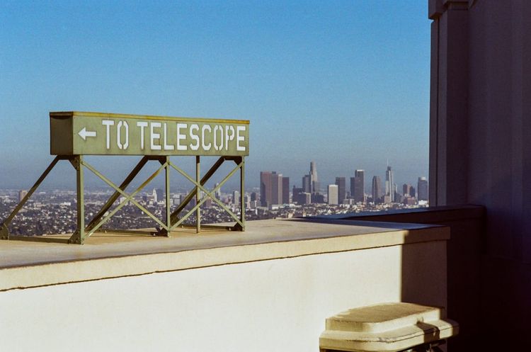 a color photograph of a metal sign at the Griffith Observatory. The sign reads "To Telescope" with an arrow pointing left. Behind and to the right of the sign is the skyline of Downtown LA