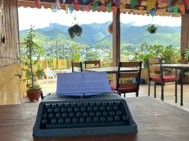a color photo showing a portable typewriter on a table in the foreground, with an open-air patio in the background and green mountains in the distance