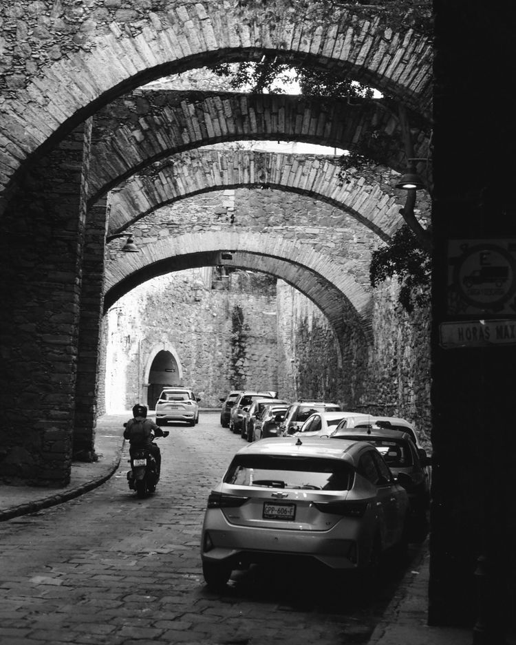 a black-and white photo of a sunken street in Guanajuato, paved with stone. cars line one side, a scooter drives on the other. The walls are stone and stone arches stretch over top.