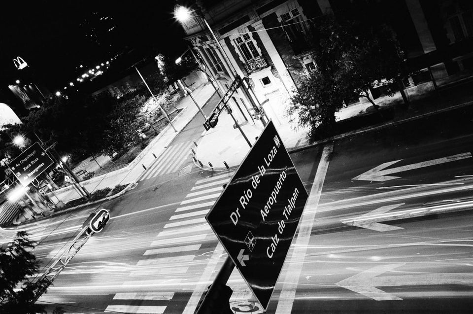 a black and white photo of a street in Mexico City from above. A street sign in the foreground, light streaks from passing cars along the roadway