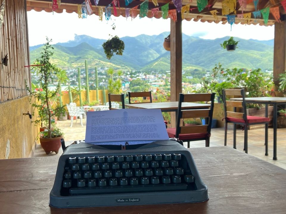a color photo showing a portable typewriter on a table in the foreground, with an open-air patio in the background and green mountains in the distance