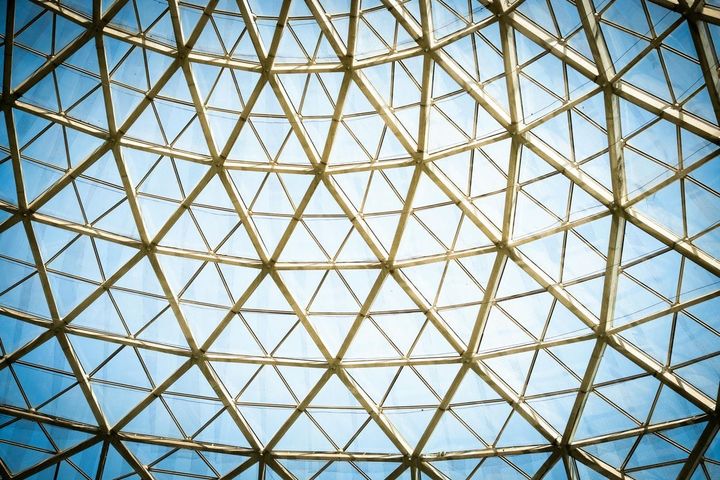 Geometric glass dome ceiling with triangular steel framework viewed from below against a blue sky, representing structured architectural frameworks in enterprise procurement.