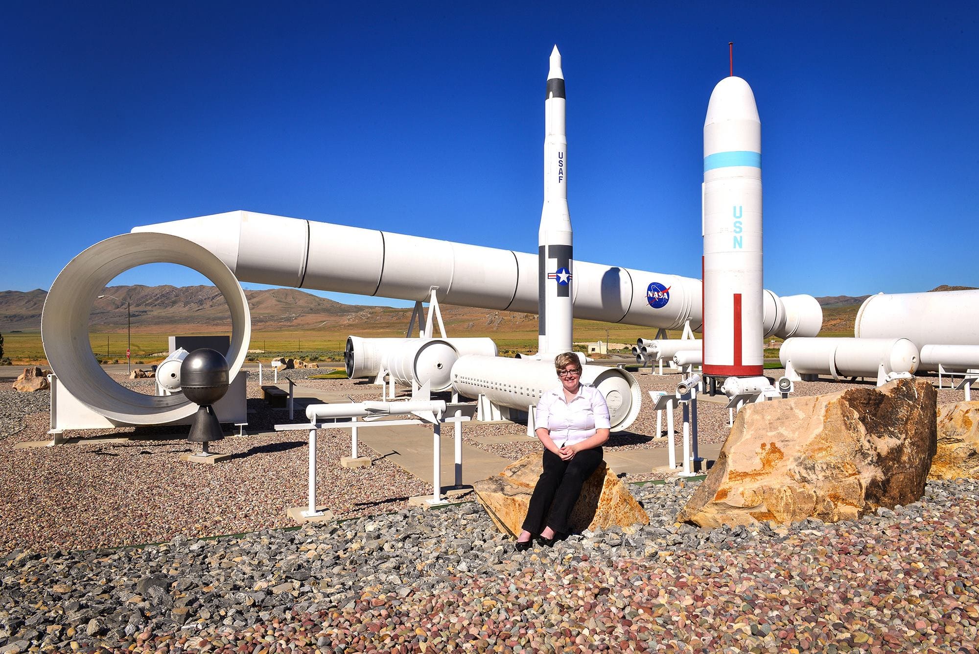 A pale-skinned blonde femme sits on a rock in front of a static display of various rockets and missiles. She is suffering under the desert heat but her smile is genuine.
