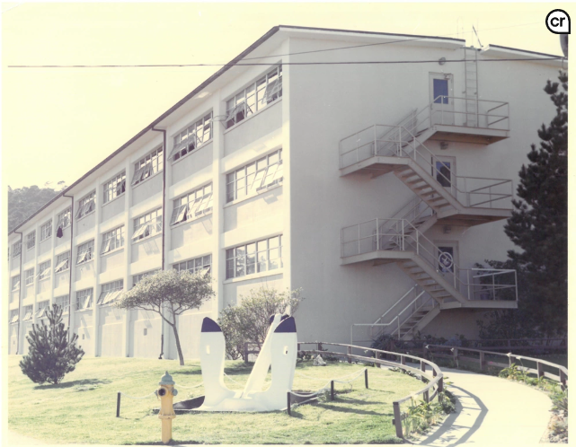 An aged photo of a three-story cinder-block building. In the foreground is a lawn with a few trees, a carefully-mowed lawn, a fire hydrant, and a decorative ship's anchor.