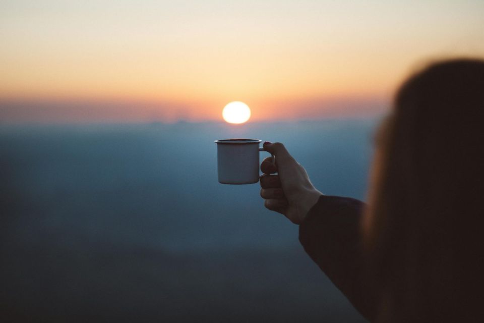 A person holding a white coffee cup out just below the sun, which is rising over a hazy, out-of-focus horizon.