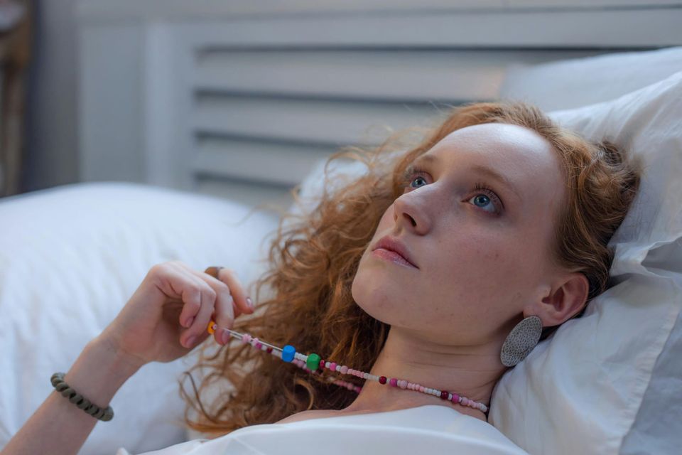 A woman lies in a soft bed, pulling thoughtfully on the beaded necklace around her neck as she gazes towards the ceiling.