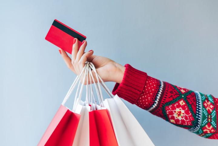 La mano de una mujer con un suéter navideño sosteniendo muchas bolsas de compras festivas y una tarjeta de crédito roja.