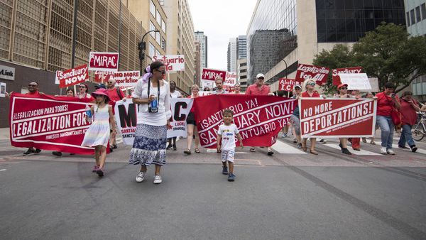 Demonstrators walk down a city street. Red and white banners read Legalization for All, Sanctuary Now! and No More Deportations