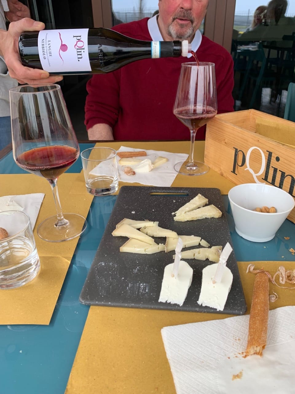 A hand pours a bottle of red wine into a glass. There are cheeses and hazelnuts on the table