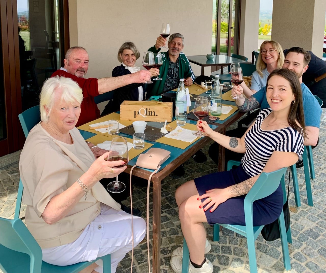 A group of people sitting at a table holding up glasses of red wine and smiling to camera.