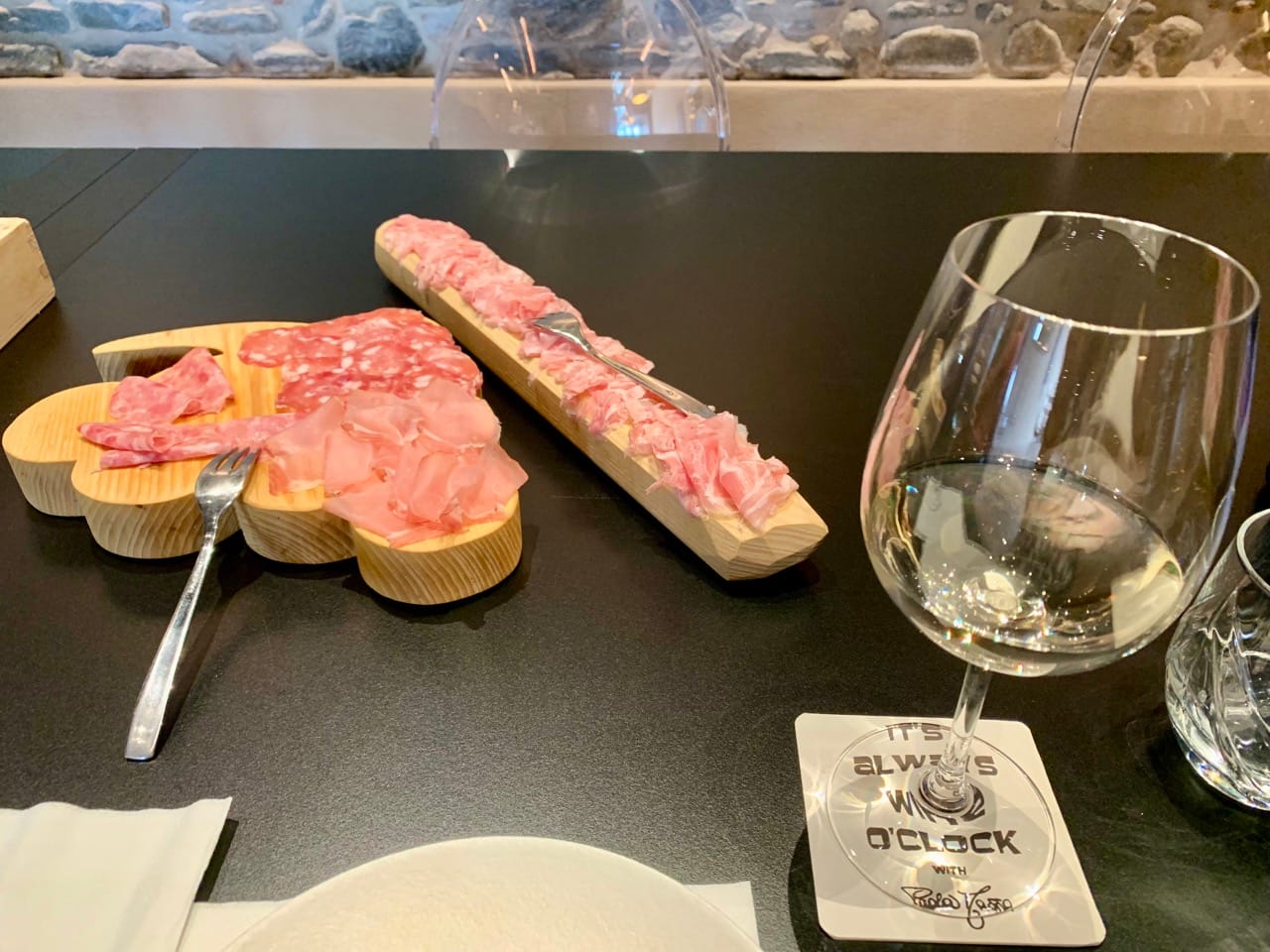 A glass of white wine with two boards of cured meat in the background.