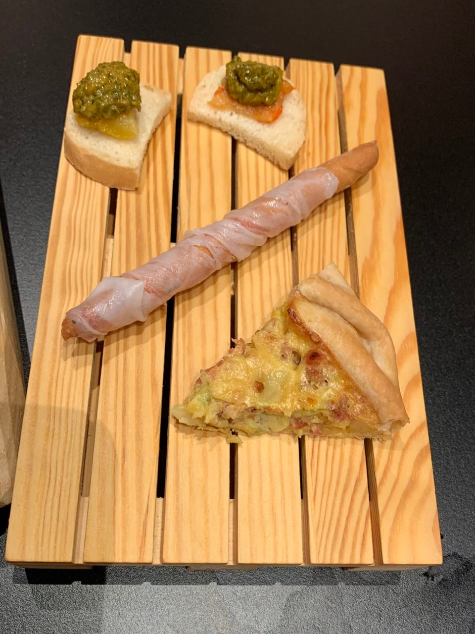 Peppers on bread with a green sauce, a breadstick wrapped in lard, and a quiche on a wooden board