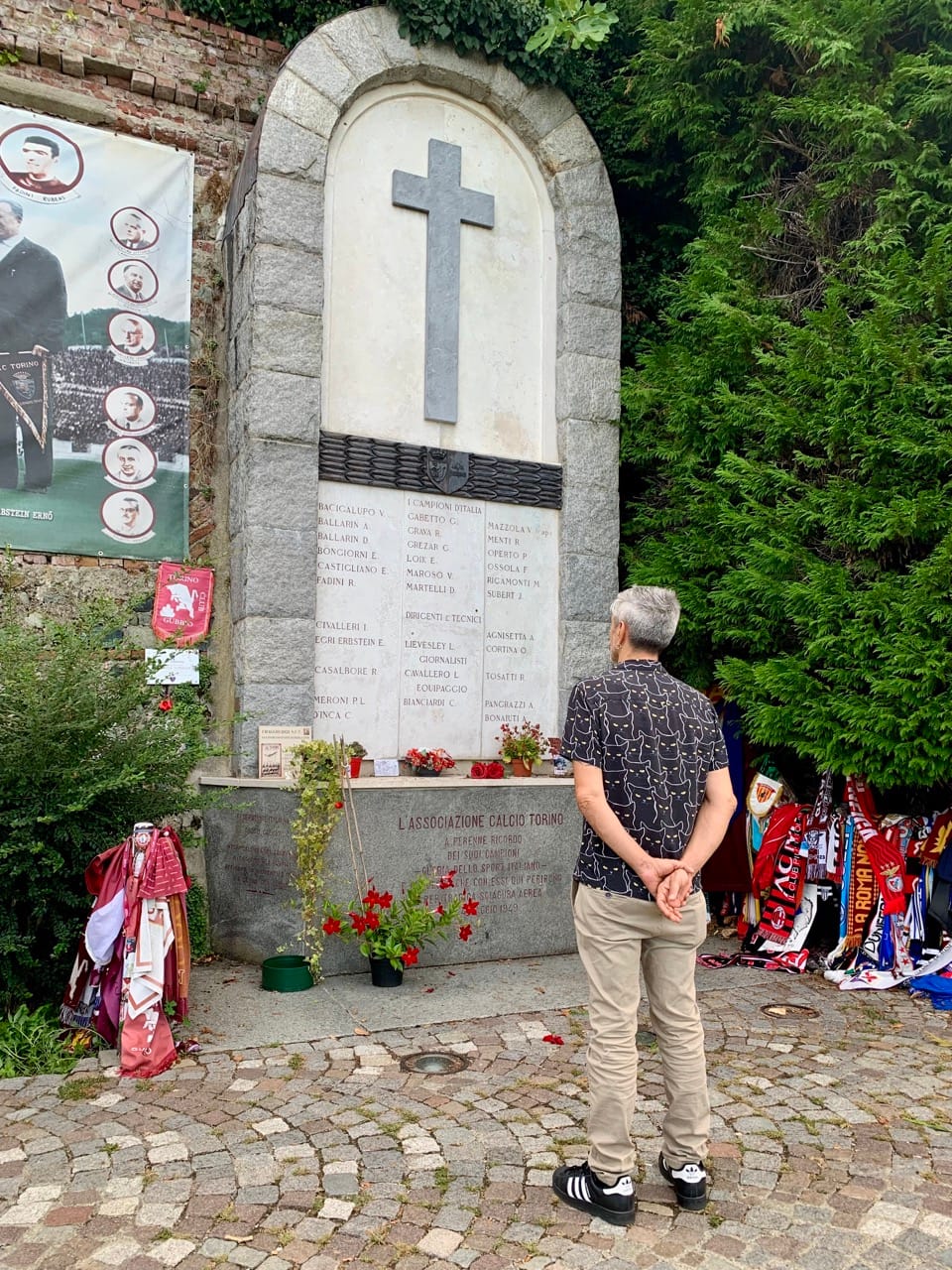 A man in a black shirt and beige trousers stands in front of a memorial surrounded by football scarves.