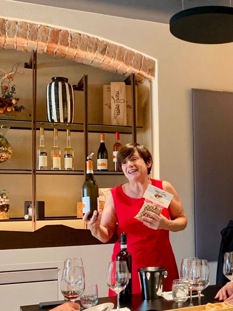 A woman in a red dress holds up a wine bottle and packet of hazelnuts in front of a table full of wine glasses