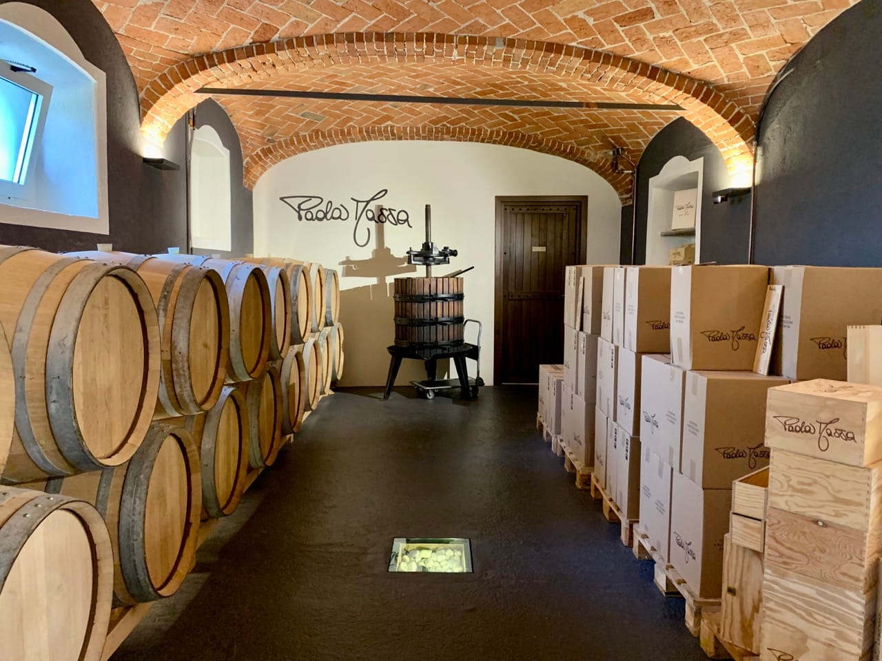 A cellar with vaulted brick ceiling, barrels and boxes lining the walls, and a grape press.