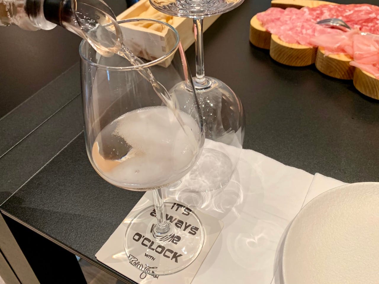 Pink spumante being poured into a glass with cured meats in the background. The coaster says "It's always wine o'clock with Paola Massa"