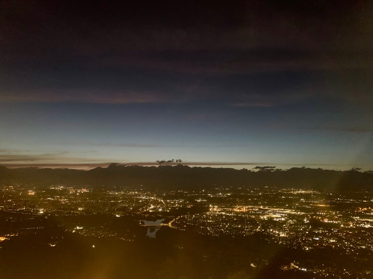 A city seen from above at night, with lights twinkling and mountains in the background lit by the last light of evening.