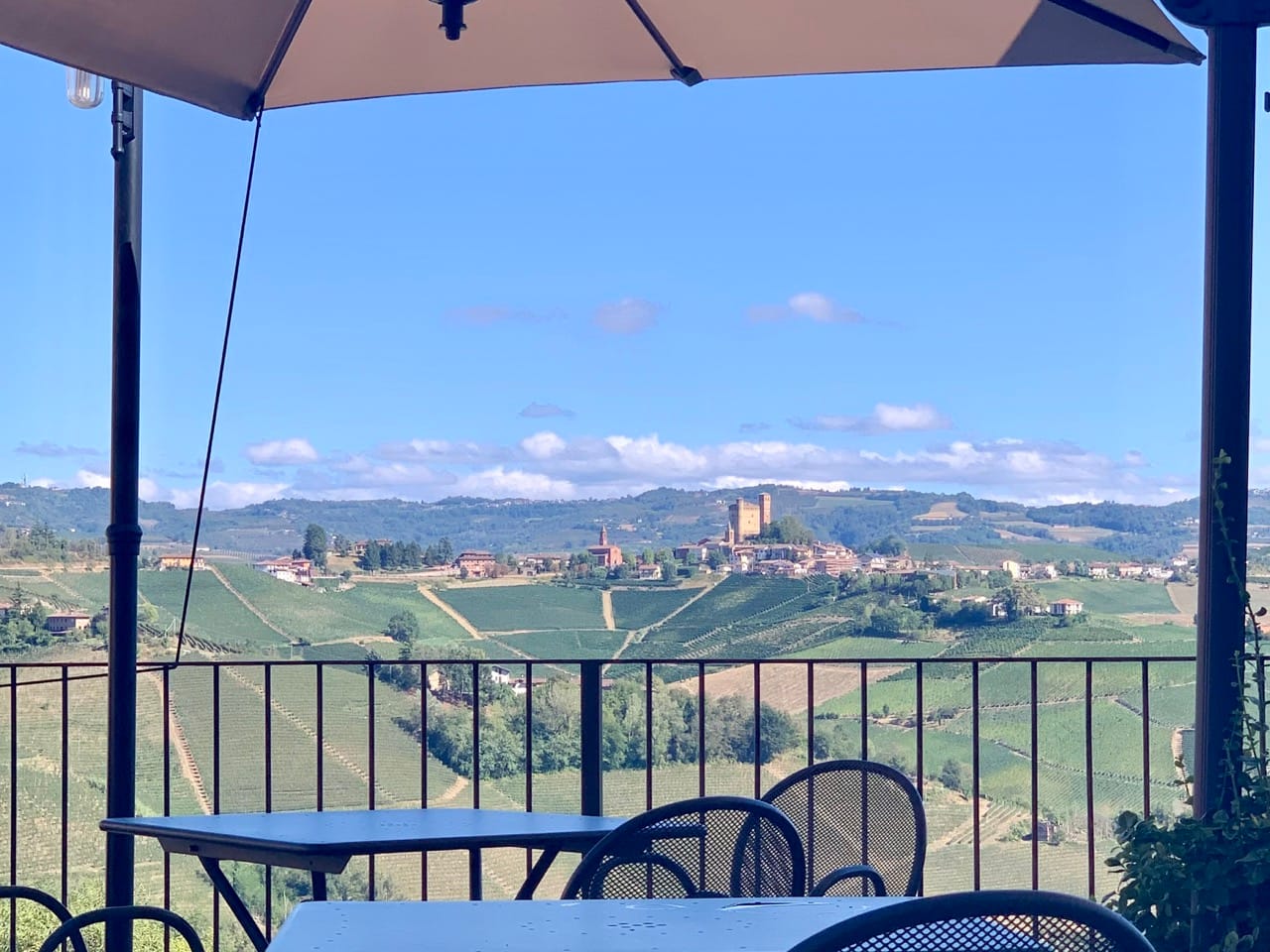 Tables and chairs on a terrace overlooking a sunny hillside with vineyards and a castle.