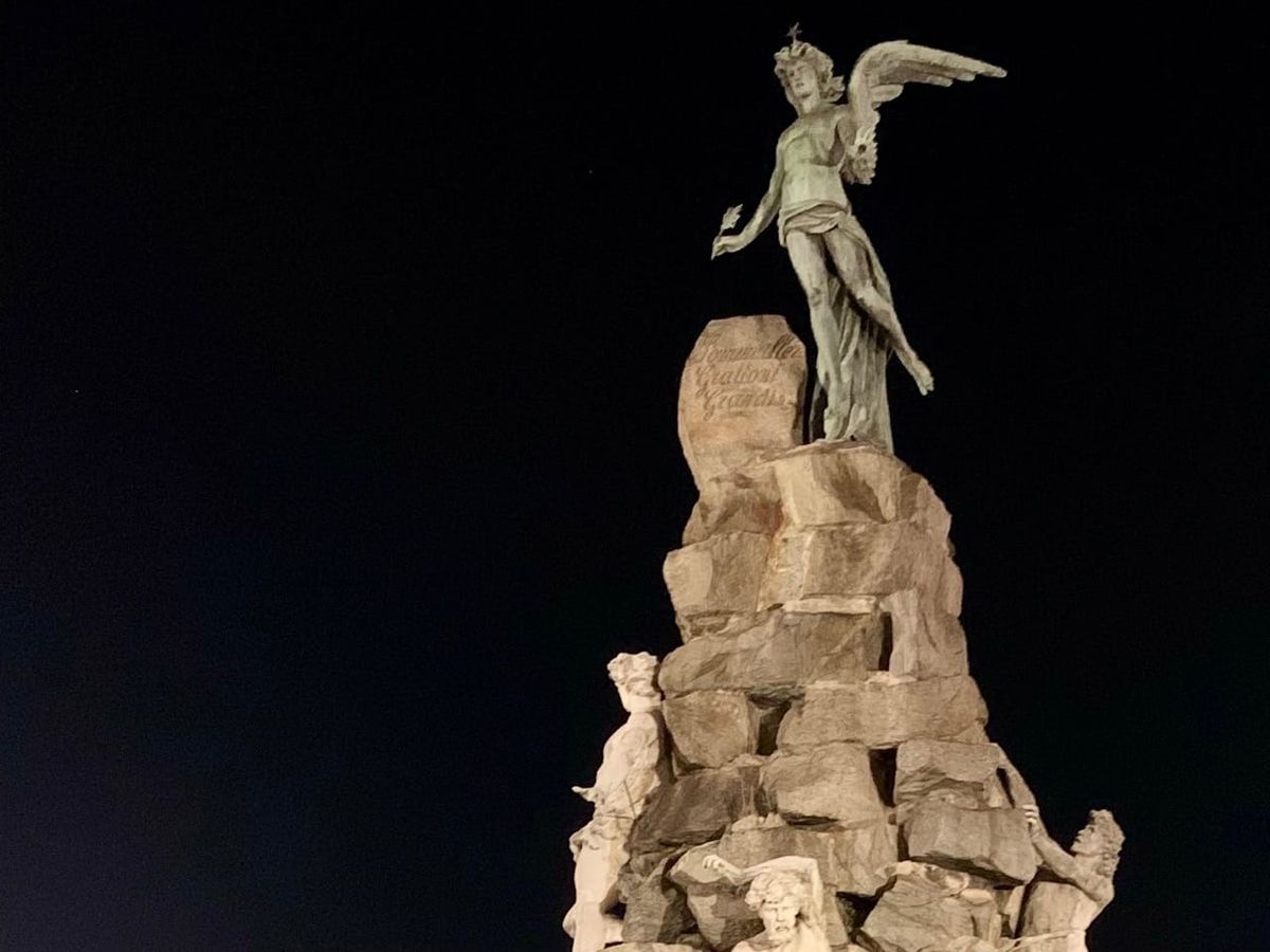A statue of a winged figure atop a pile of stones holding a quill in his right hand and his left hand palm facing down
