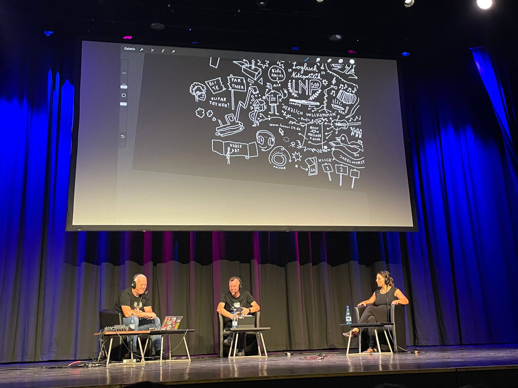 Eine große Bühne mit drei sitzenden Menschen: Tim Pritlovem Linus Neumann und Elena Gußmann, im Hintergrund graphic recording, weiße Linien auf schwarzem Grund
