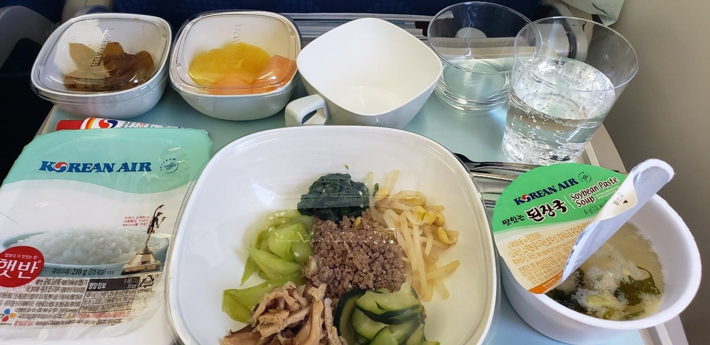 Plane food on Korea air. Different dishes, including bibimbap, rice, fruit, and miso soup, along with three cups.