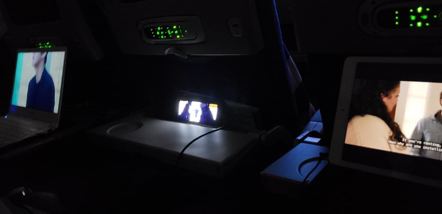 Three screens playing media in the dark on an airline flight. Laptop on the left, phone in the middle, iPad on the right.