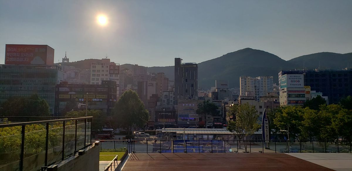 A cityscape in Busan, South Korea, with various size buildings. Large hills can be seen rising behind the buildings. The sun shines.