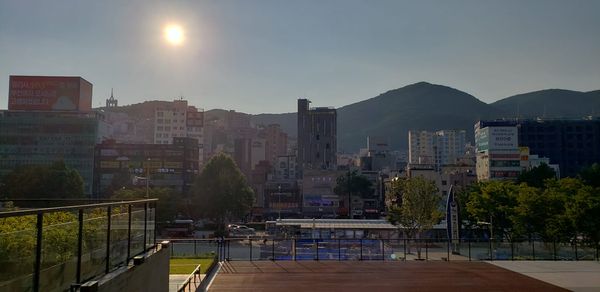 A cityscape in Busan, South Korea, with various size buildings. Large hills can be seen rising behind the buildings. The sun shines.