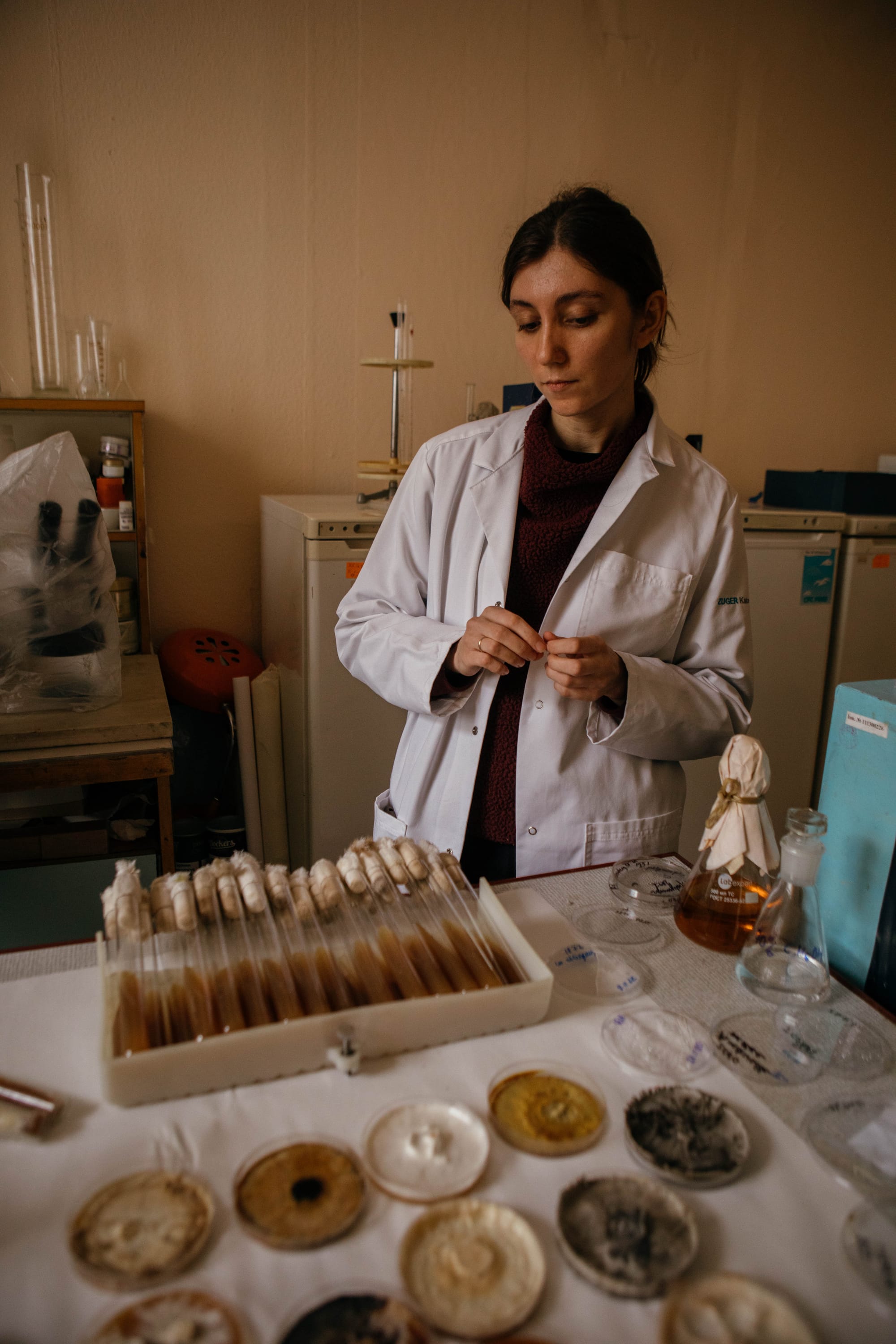 Alisa Atamanchuk, wearing a white lab coat, checks out test tubes containing different fungal strains in the lab. She looks preoccupied.