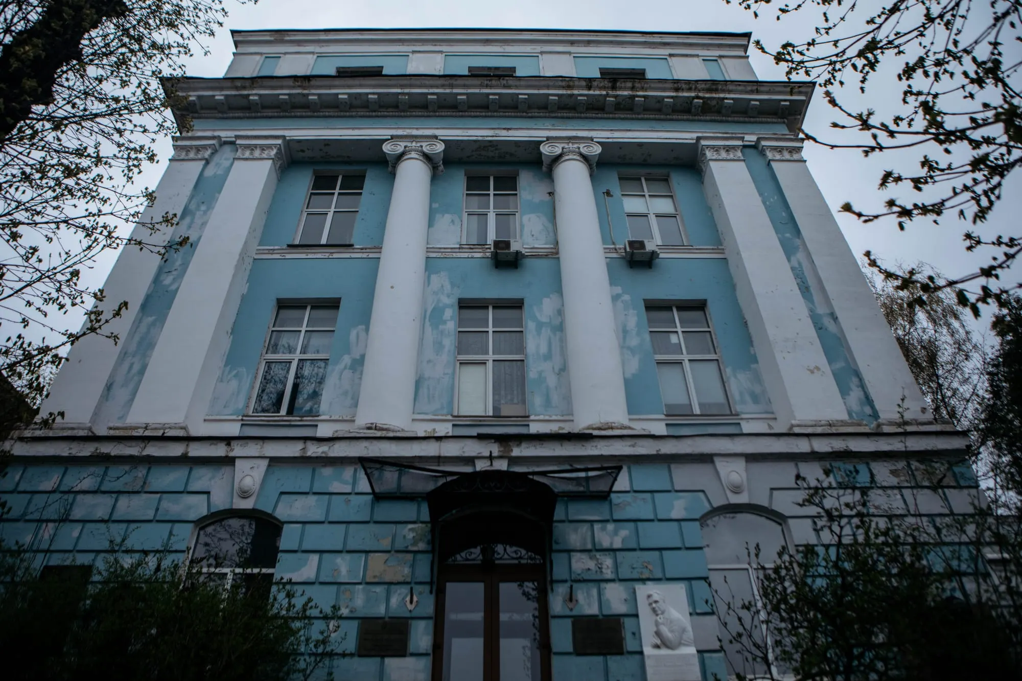 A light blue-and-white building, with colonial-era architecture, stands amid trees and foliage.