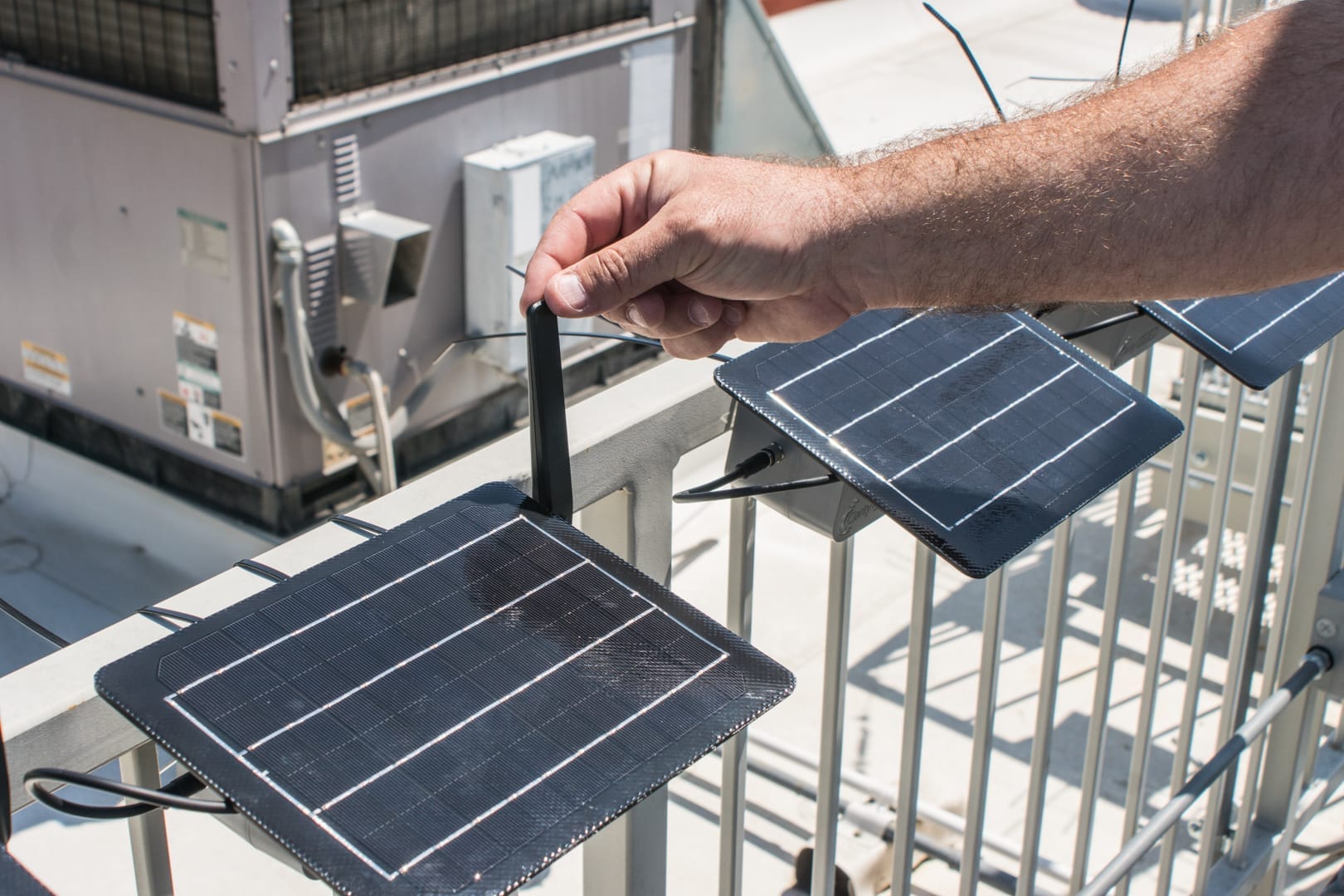 A person is seen adjusting an antenna on a small solar-powered monitoring device mounted on a rooftop railing.