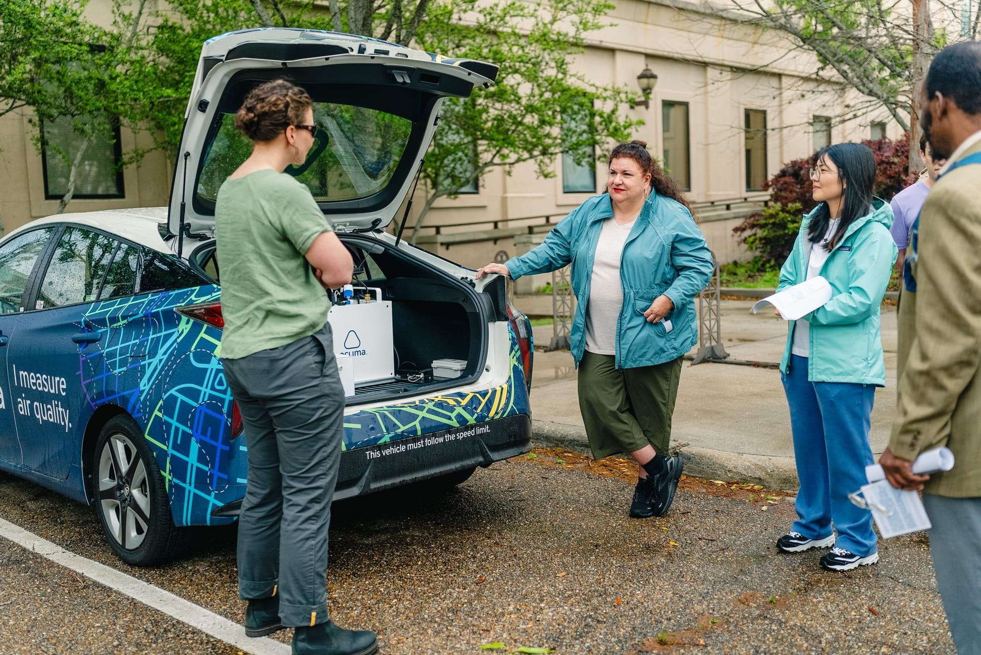 A small group is hanging out by the open trunk of a vehicle wrapped with "I measure air quality" graphics. They’re inspecting air-testing equipment loaded in the back.
