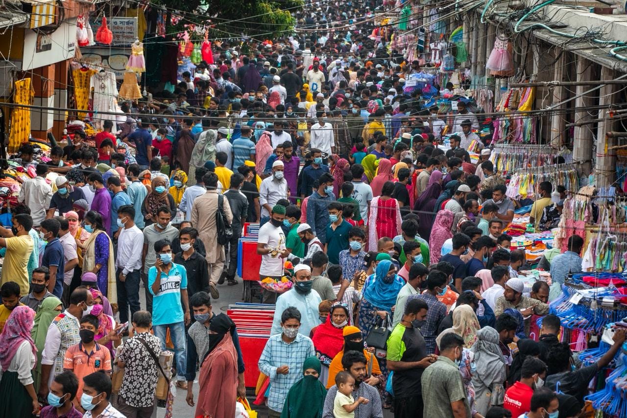 A crowded market street lined with clothing shops on both sides. Most people are seen wearing masks and caps.