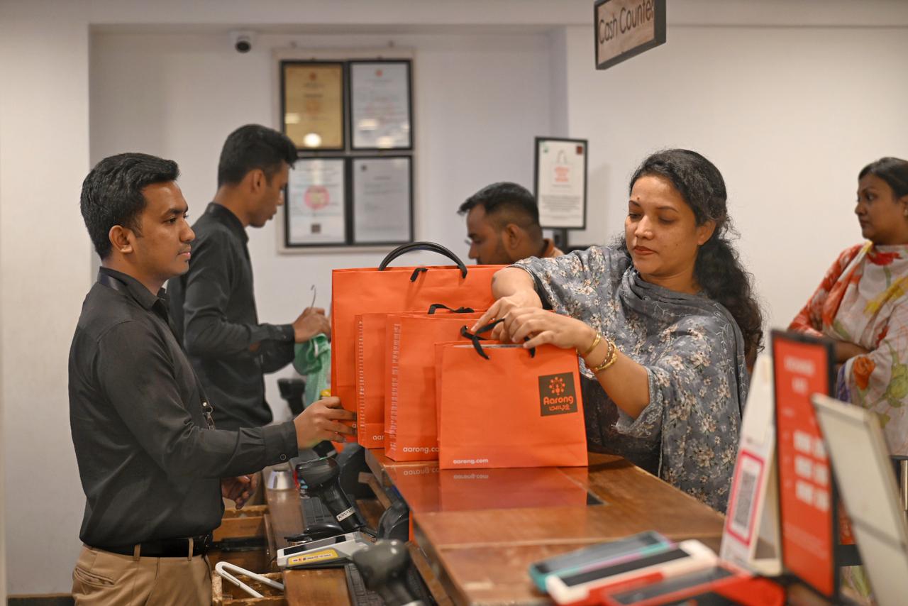 A customer holds an orange Aarong shopping bag while a shop employee in black and cream formalwear looks on.