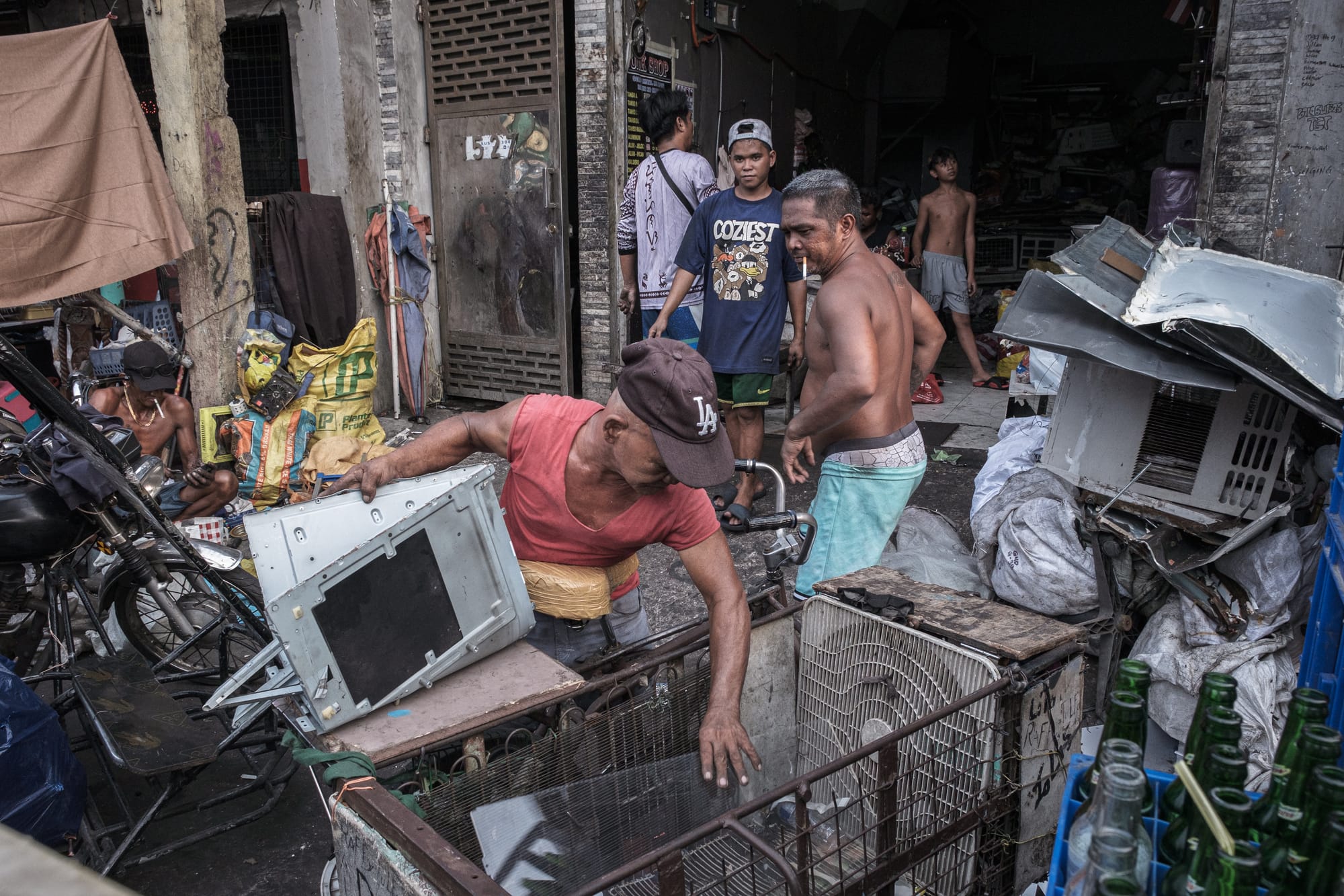 A brown man in a red shirt and a brown Los Angeles Dodgers cap picks up the scraps of an air conditioner. Behind him are other workers, and to his rights are more AC scrps and empty glass bottles.