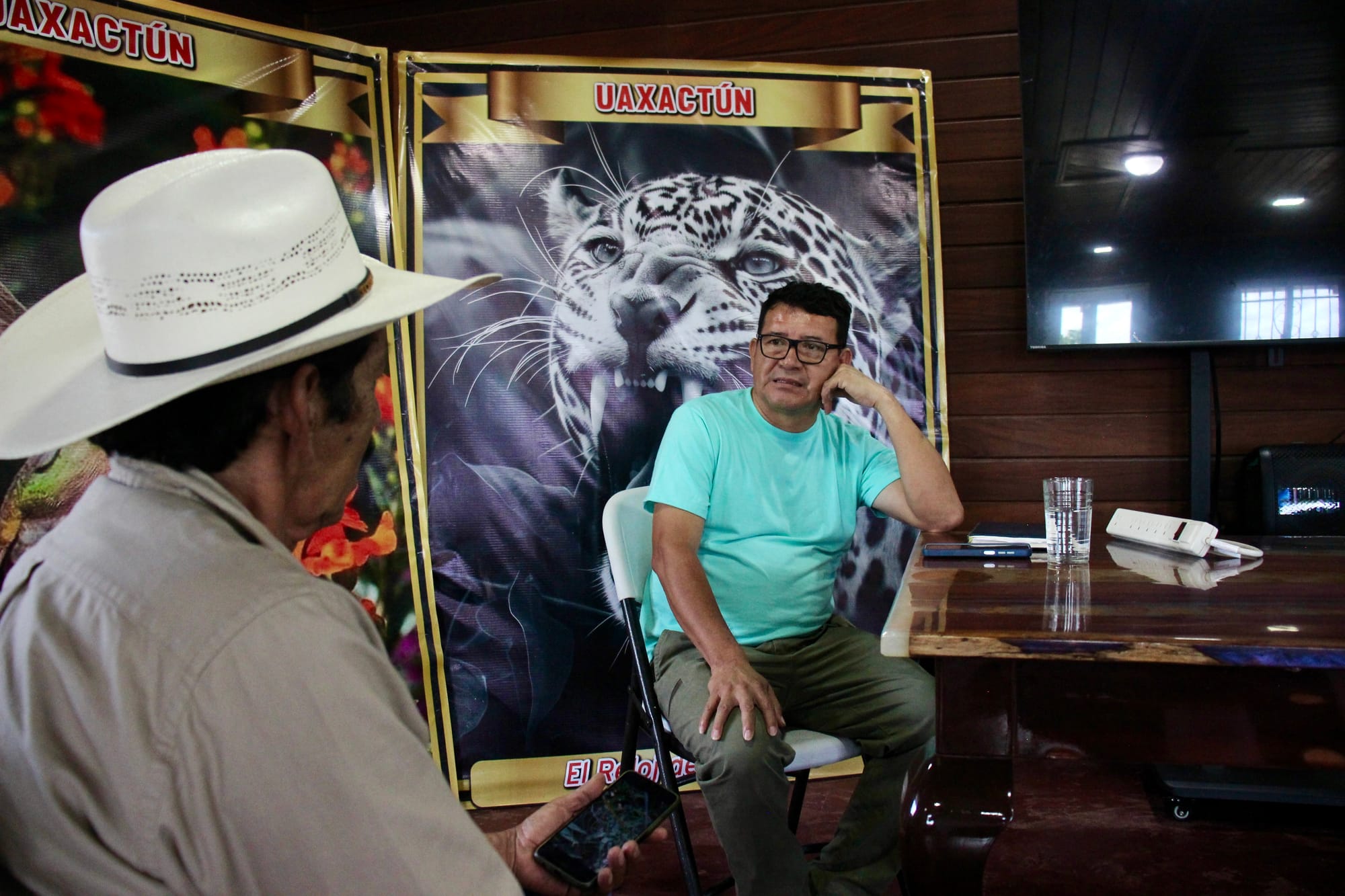 Two people sit facing each other in an office beneath a large poster reading “UAXACTÚN” with an image of a roaring lion.