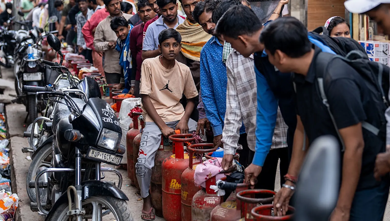 A line of people wait with LPG cylinders along a busy roadside. A boy sits among them as others hold their place.