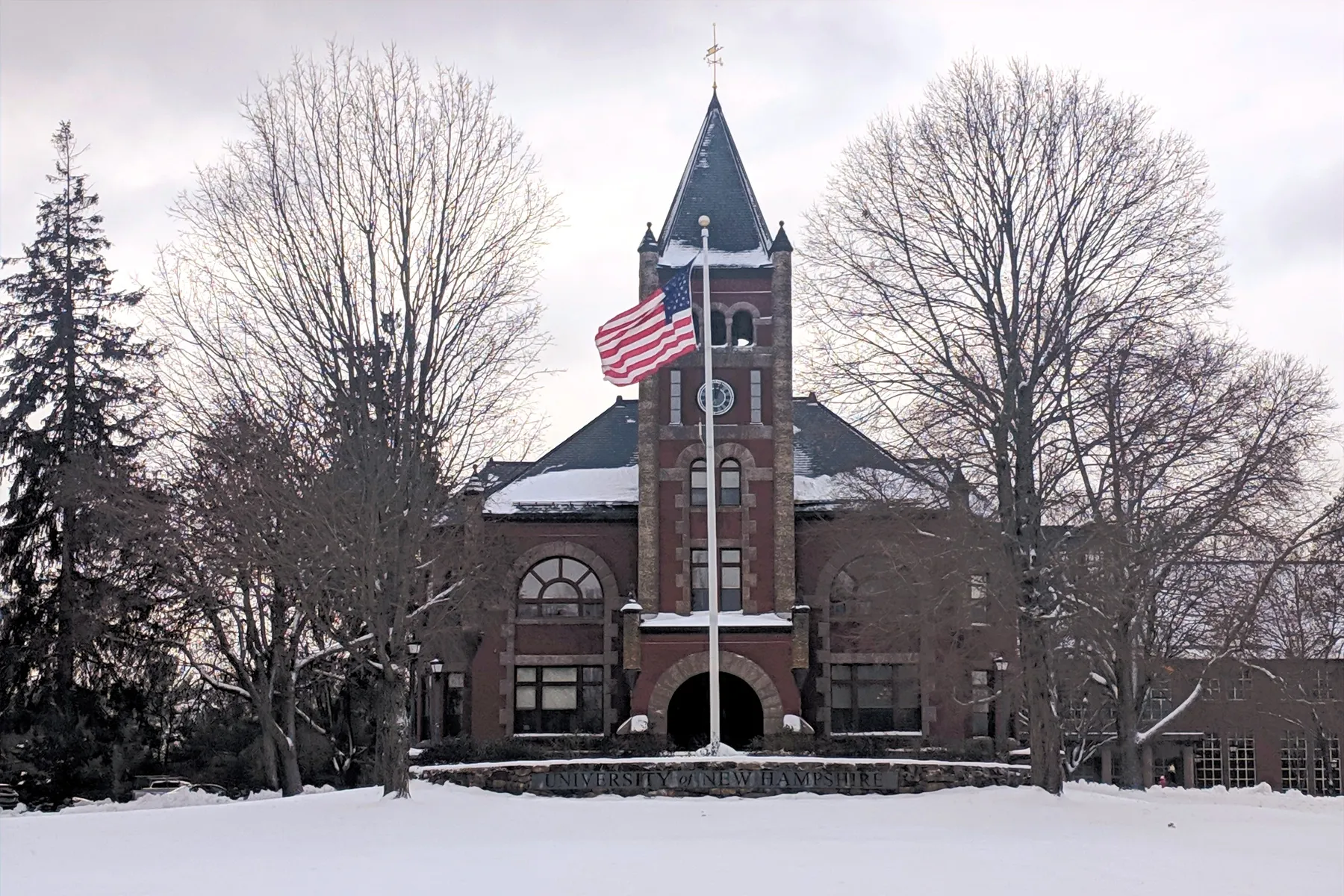 A snow-covered University of New Hampshire building stands with an American flag flying in front, surrounded by dried pine trees