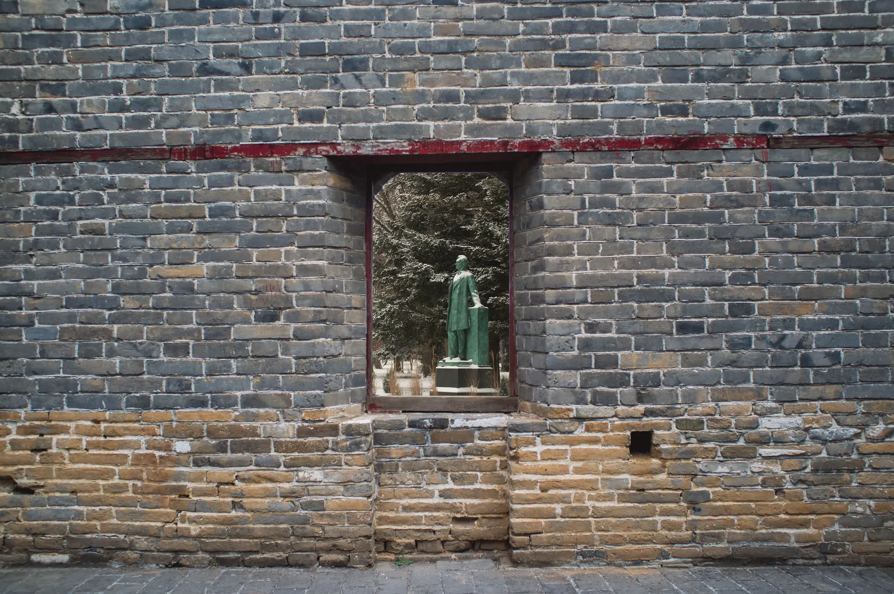 A grey brick wall with a rectangular opening which serves as an entrance. Through it, one can see a green statue surrounded by vegetation