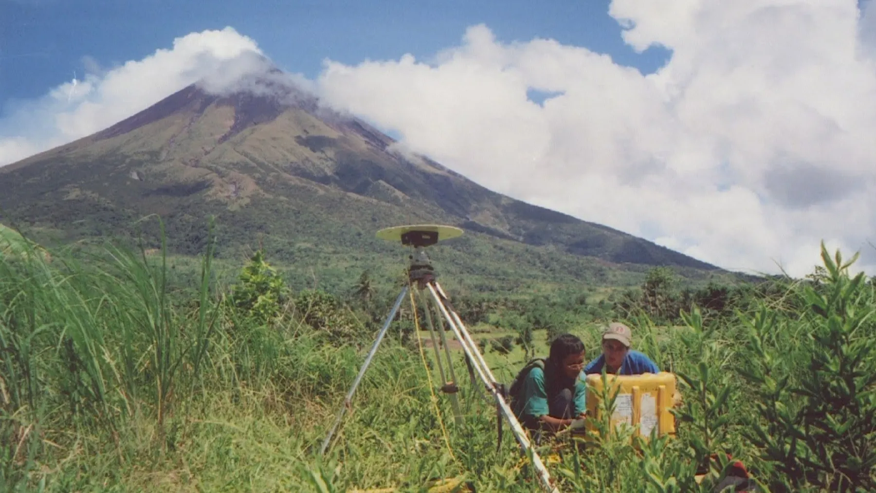 A cloud-capped mountain peak in the background. Two researchers work in the field next to a tripod-mounted setup.